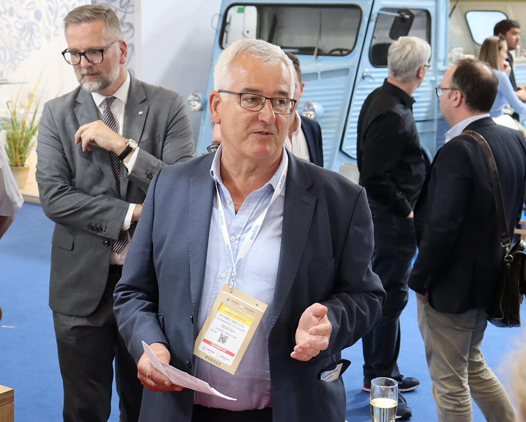 Photo of a man at an event in the middle of a conversation. There are other people having conversations in the background.