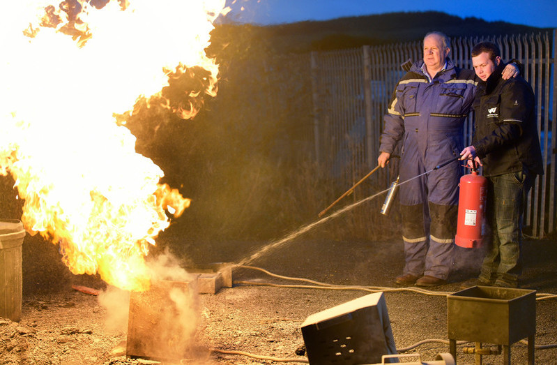 A trainee is putting out a fire with the trainer standing beside him for safety